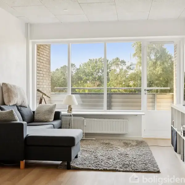 Sofa står stille i stuen, vendt mod et stort vindue, der lukker naturligt lys ind, flankeret af en reol med et TV. Udenfor ses træer.