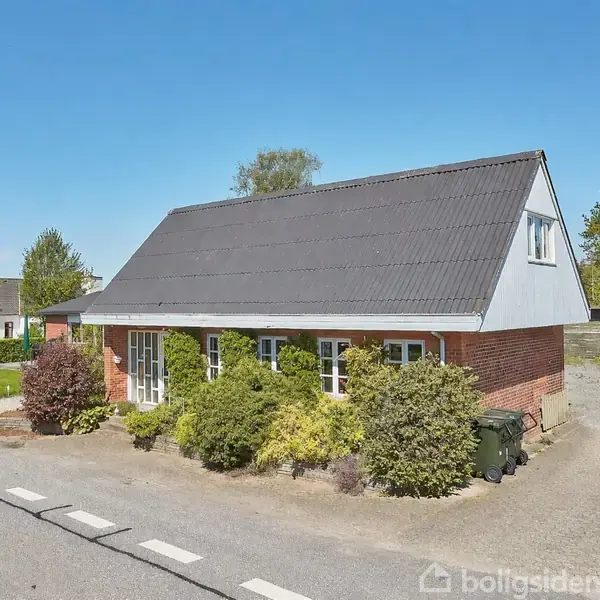En muret bungalow med høj tagbeklædning står ved siden af en vej. Huset er omgivet af buske og har en indkørsel til højre.