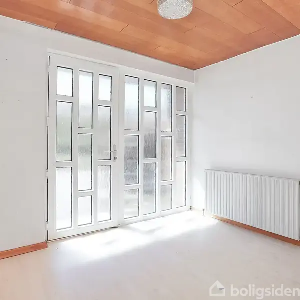 Et lyst rum med et geometrisk mønstret glasparti. Sollys strømmer ind. Væggene er hvide, og der er en radiator under vinduet. Loftet er træbeklædt.