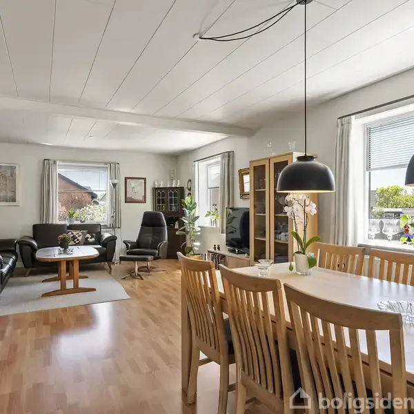 Spisebord med træstole står i forgrunden, mens en sofagruppe med sorte lædermøbler ses i baggrunden. Rummet har lyse vægge og store vinduer, der giver naturligt lys.