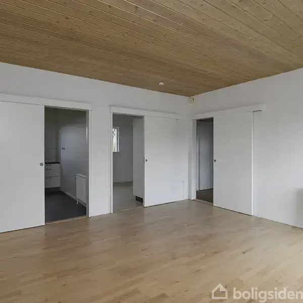 Et tomt rum med lyse trægulve og loft, tre skydedøre der fører til tilstødende rum, og en radiator på væggen. Væggene er hvide, hvilket skaber en minimalistisk atmosfære.