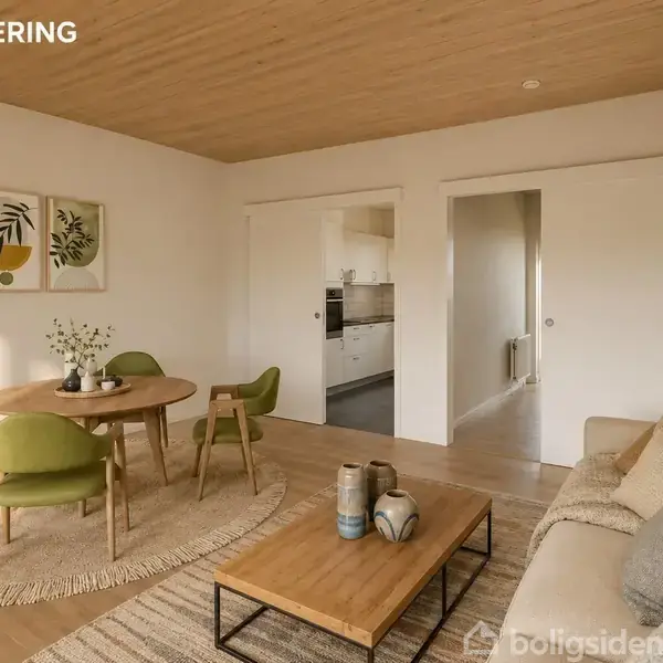 Stue indrettet med en beige sofa, firkantet træbord og runde spisebord omgivet af grønne stole. Køkken ses gennem en døråbning. To billeder hænger på væggen. Tekst: "VISUALISERING".