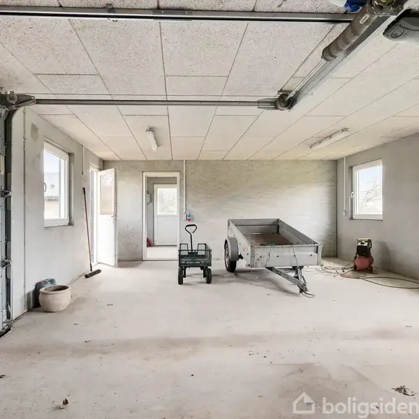 En garage indeholder en åben trailer og en trallevogn. Rummet har en stor port til venstre, to vinduer og en dør til højre. Loftet har rør og lysarmaturer.
