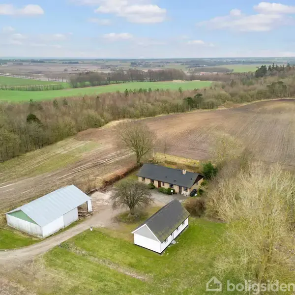 Luftfoto af landsted med tre bygninger: en hvid bygning, et hus med mørkt tag, og en lade. Omgivet af marker og skovbevoksning under en let overskyet himmel.