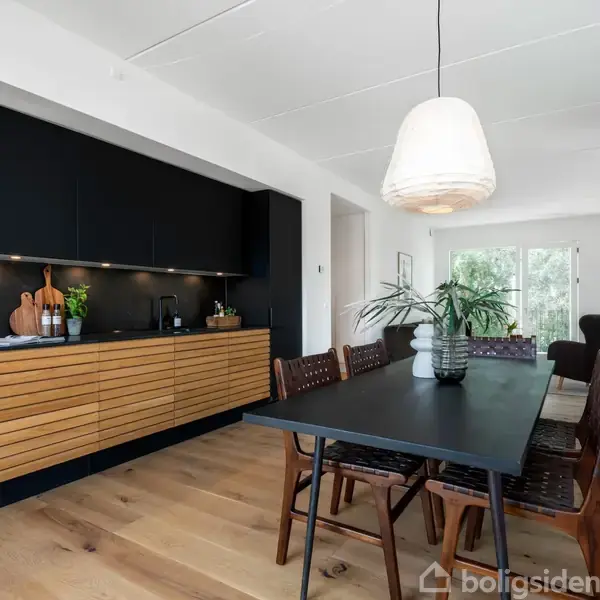 Et spisebord med seks stole står i en moderne spisestue. En stor lampe hænger ovenover. Til venstre ses et køkken med træskabe og mørke fronter.