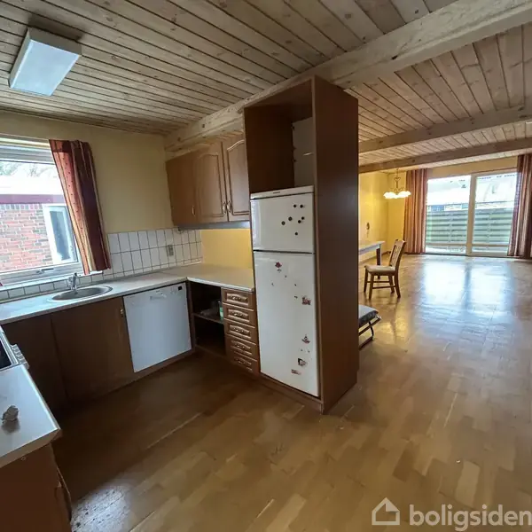 Køkkenet har hvidevarer og træskuffer, åbner sig mod en spisestue med træbord. Store vinduer giver lys, mens trægulve og loft skaber en hyggelig atmosfære.