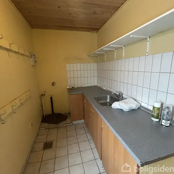 Et lille rum med en gul væg og flisegulv har en bordplade med vask langs den ene væg. Hylder er monteret over bordpladen, og kroge langs den modsatte væg. Ingen tekst er synlig.