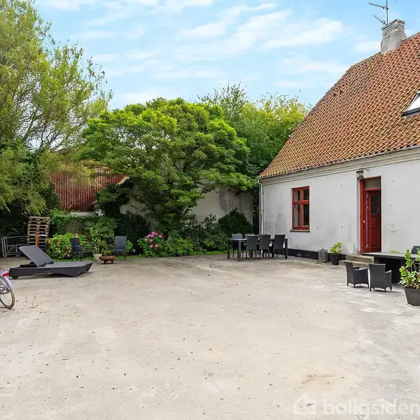 Et hus med rødt tag står ved siden af en åben gårdsplads. To cykler er parkeret, og der er havemøbler blandt grønne planter og buske i baggrunden.