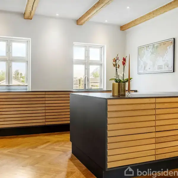 Køkken med moderne trælameller og sort køkkenø. To vinduer giver dagslys. Trægulv og synlige loftsbjælker skaber en varm atmosfære. Billede på væg og blomsterdekoration på bordet.