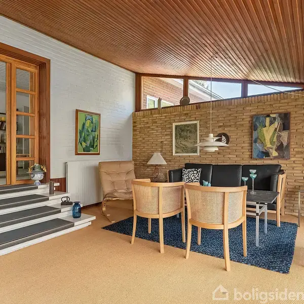 En stue med træloft og gulvtæppe, en sofa og stole omkring et lavt bord. Væggene er dekoreret med kunst, og der er store vinduer med lysindfald.