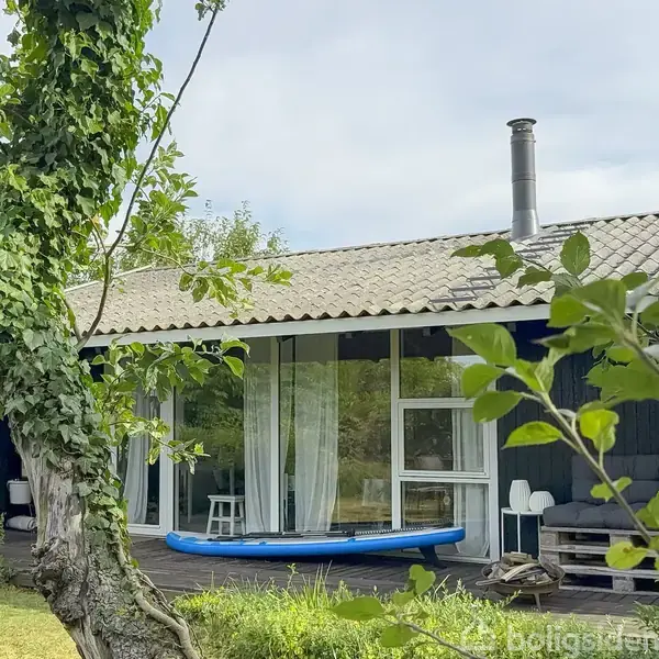 Et sort træhus med store vinduer vender mod en frodig have. En blå oppustelig kajak ligger på terrassen. Hvidt gardin synligt gennem vinduet. Grønt buskads omgiver området.