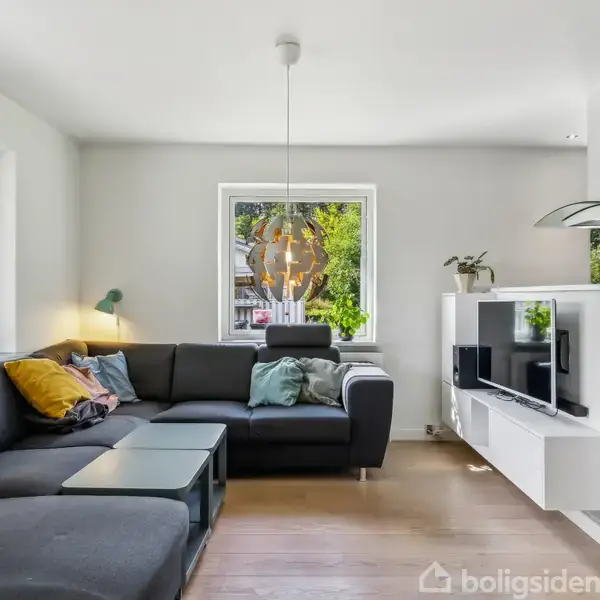 En sort hjørnesofa med farverige puder står i et lyst stueområde. Ved siden af er et glasbord, og en moderne hængelampe hænger fra loftet. Store vinduer giver udsigt til grønne træer udenfor. En tv-reol med fjernsyn og planter står ved siden af køkkenet.