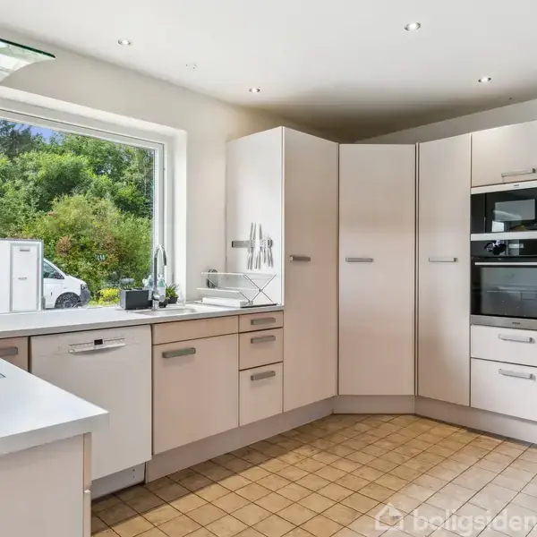 Køkken med lyse skabe og apparater, hvor et vindue giver udsigt til en grøn have. Rummet har flisegulv og indbyggede ovne.