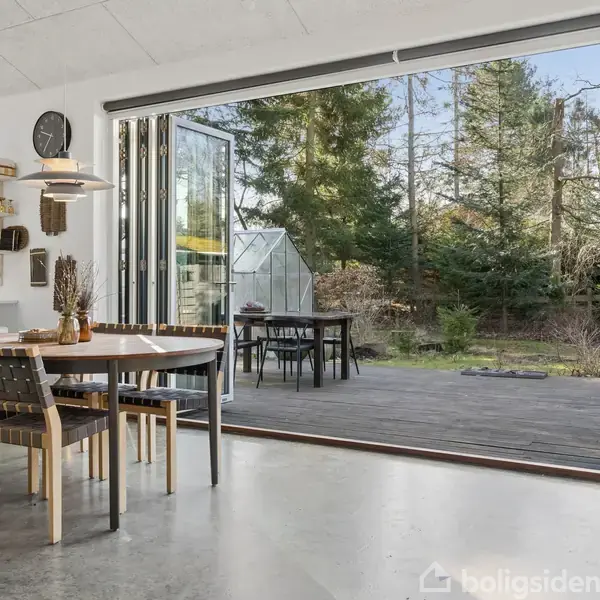 Et åbent spiseområde med et rundt bord og fire stole ved siden af skydedøre, der fører ud til en træterrasse omgivet af en skov fyldt med høje træer.