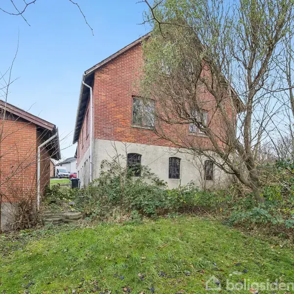 Rødt murstenshus med skråt tag står ved siden af en mindre bygning, omgivet af græs, buske og træer. En smal stentrappe leder op mellem bygningerne.