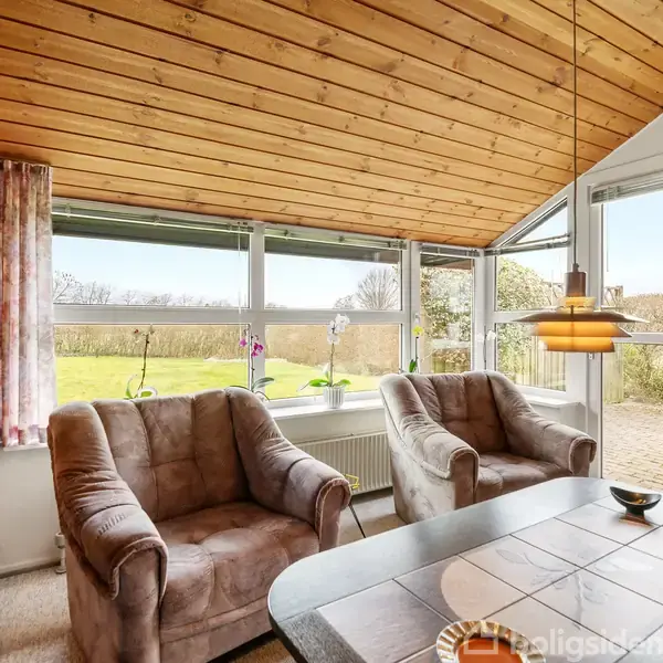 To lænestole står foran store vinduer med udsigt til en have. Loftet er træbeklædt, og ved siden af en bordlampe hænger over et spisebord. Gardinerne er lyserøde.