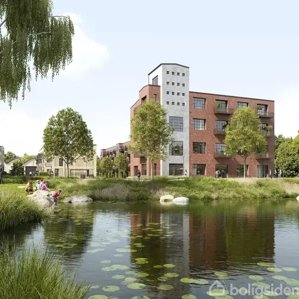 Bygning med røde mursten står ved siden af en sø med åkander, omgivet af træer og græs. Mennesker sidder og nyder omgivelserne ved vandkanten.