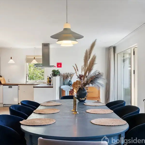 Spisebord med syv blå stole er pyntet med bordskånere og en vase med tørrede blomster. Det står i et lyst køkkenalrum med hvide skabe og store vinduer.