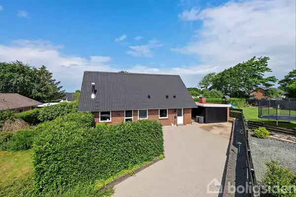 Et rødt murstenshus med sort tag står i en grøn have med buske. Haven har en trampolin og en lys belægning foran huset under en blå himmel.