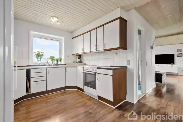 Køkken med hvide og brune skabe, en ovn og vask. Blomster på bordet ved vinduet. Trægulv og åben forbindelse til stue.