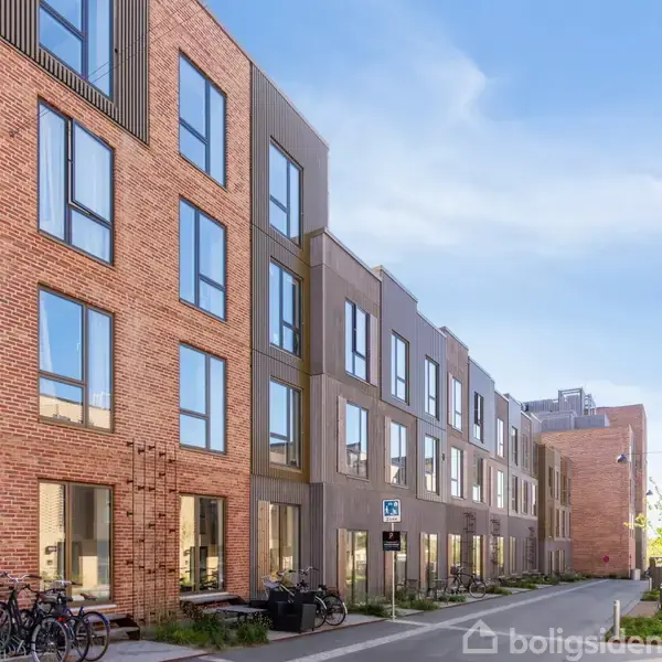 Moderne lejlighedsbygning strækker sig langs en stille gade med cykler parkeret foran. Sollys reflekterer i store vinduer. Grønne planter kantet langs fortovet giver en rolig atmosfære.
