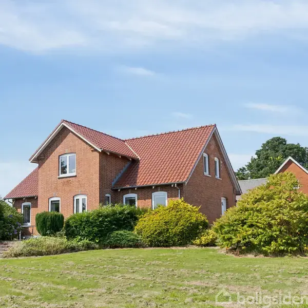 Et rødt murstenshus står i en frodig have med grønne buske og træer. Huset har et rødt tegltag og hvide vinduer. Blå himmel og træer i baggrunden.