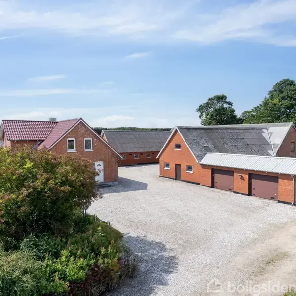 Et rødt murstenshus med to tilhørende bygninger står forbundet i en gårdsplads med grus. Omkringliggende grønne buske og træer danner en landlig baggrund under en blå himmel.