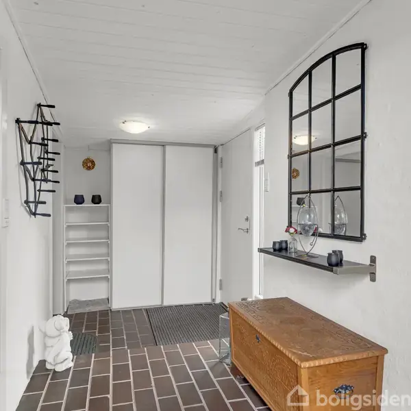 En entré med et træloft og murstensgulv har en træbænk og en hylde med glasgenstande. Et spejl hænger på væggen. Der er et skab og nogle hylder i baggrunden.