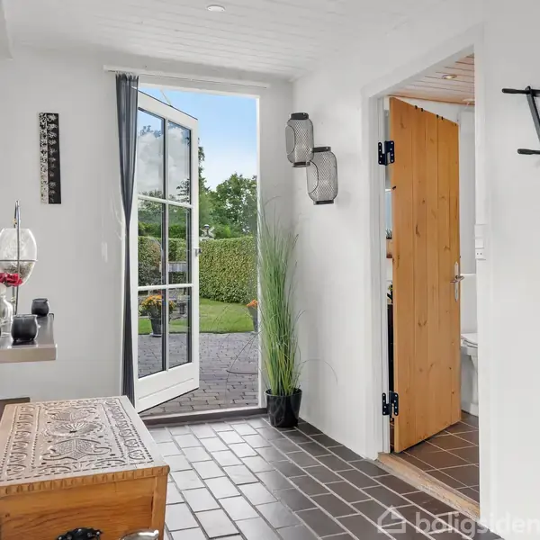 Dør med glas åbner mod en have, hvor grønne buske ses. Indendørs er gulvet mørkt flisebelagt og dekoreret med en udskåret træbænk og sort vægpynt.