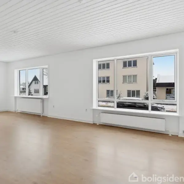 Et tomt rum med lyse trægulve har store vinduer, der viser en udsigt til huse og snebeklædte omgivelser udenfor. Væggene er hvide, og loftet har smalle paneler.