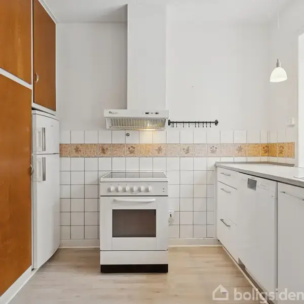 Komfur står i et køkken med hvide skabe på højre side og brune skabe til venstre. En emhætte er over komfuret. Et vindue på højre side giver naturligt lys.