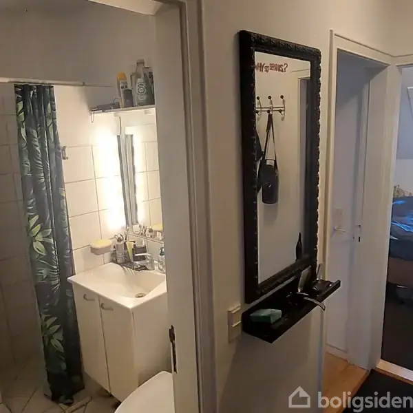 Et badeværelse med et brus med grønne gardiner bag en dør, et spejl over en vask med lys. Til højre ses en gang og et oplyst soveværelse med seng.