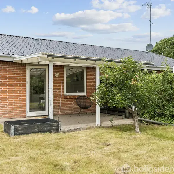 En murstensvilla med fladt tag har en lille overdækket terrasse med en stol og en plantekasse. I forgrunden ses en græsplæne og et kort træ.