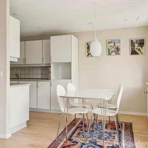 En moderne spisestue med et hvidt bord og fire stole står på et farverigt tæppe. Køkkenet med hvide skabe ses i baggrunden, og tre små billeder hænger på væggen.