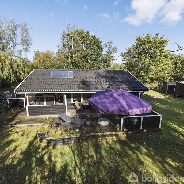Et enkelt hus med sort tag og en stor terrasse med en lilla parasol. Omgivet af en grøn have med træer og buske.