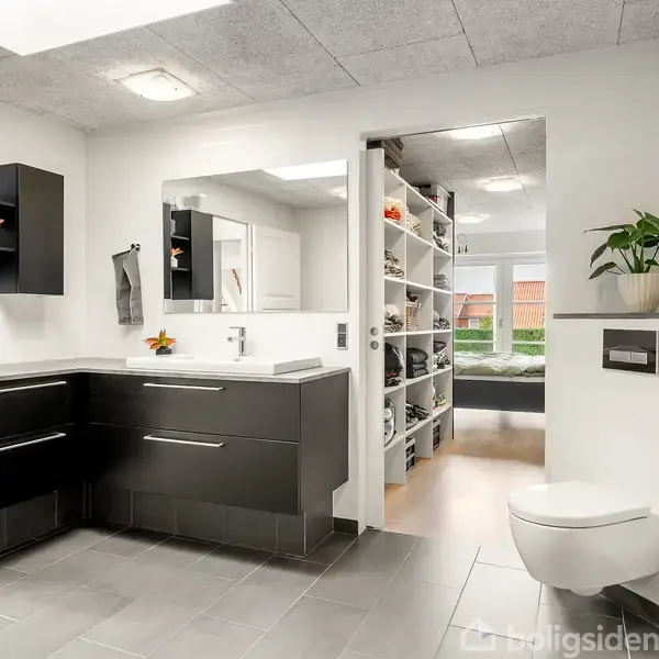 Et moderne køkken har sorte skabe og en køkkenø med frugt på. En vægmonteret tv-skærm og en plante er til stede. I baggrunden ses et spiseområde.