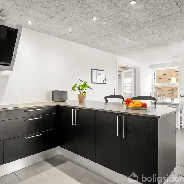 Køkken med sorte skabslåger, hvor en skærm hænger på væggen. Et bord er dekoreret med frugt og en potteplante. Rummet er lyst med spiseplads i baggrunden.