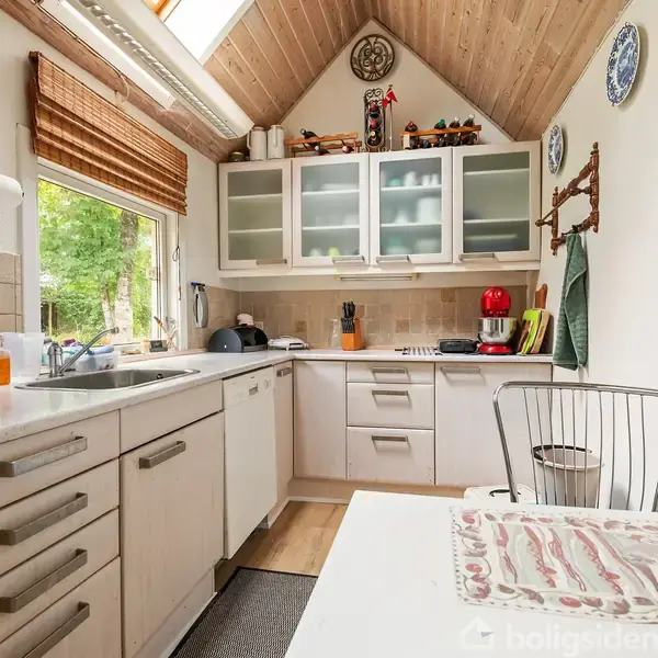 Et lyst køkken med hvide skabe og glasfronter. Der er en vask ved et vindue til venstre, og en bordplade med forskellige apparater til højre. Rummet har skrå loft og dekorative tallerkener.