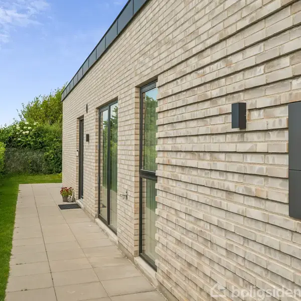 Et moderne murstenshus står stille i solen, omgivet af græs og hække. To store vinduer vender mod en flisebelagt sti, der strækker sig langs huset.