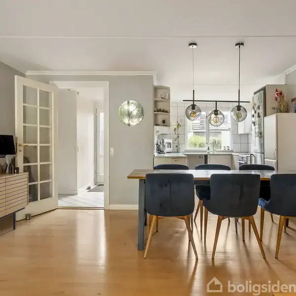 Spisebord med blå stole står i et moderne spisestue/køkkenområde. Bordet er oplyst af tre hængelamper, omkranset af træmøbler, spejle på væggen og grå vægge.