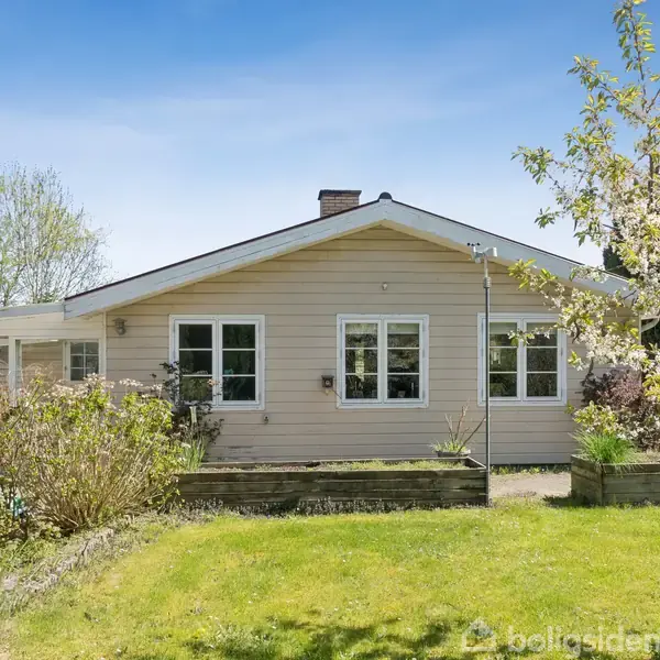 En enkeltstående villa står stille omgivet af en frodig have med grønne buske og træer. Huset har store vinduer og en skråtag, mens himlen er klar og blå i baggrunden.
