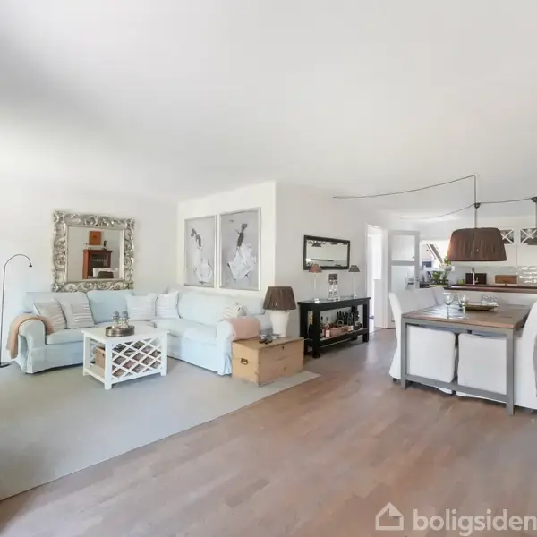 Stue med lys sofa og sofabord i hvidt træ. Til højre står spisebord og stole under en lampe, i et moderne indrettet køkkenmiljø. Ingen tekst udover "boligsiden" vandmærke.