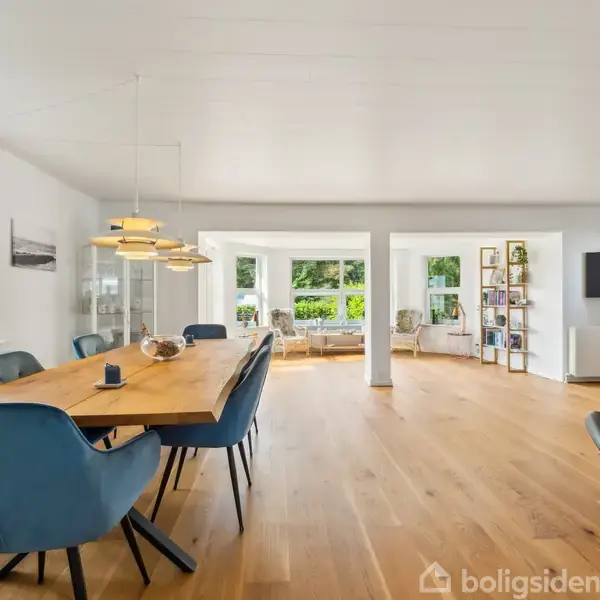 Bord med seks blå stole står i en moderne stue. Træbordet er oplyst af hængelamper, og store vinduer giver udsigt til en have. Væggene er hvide med en hylde og tv.