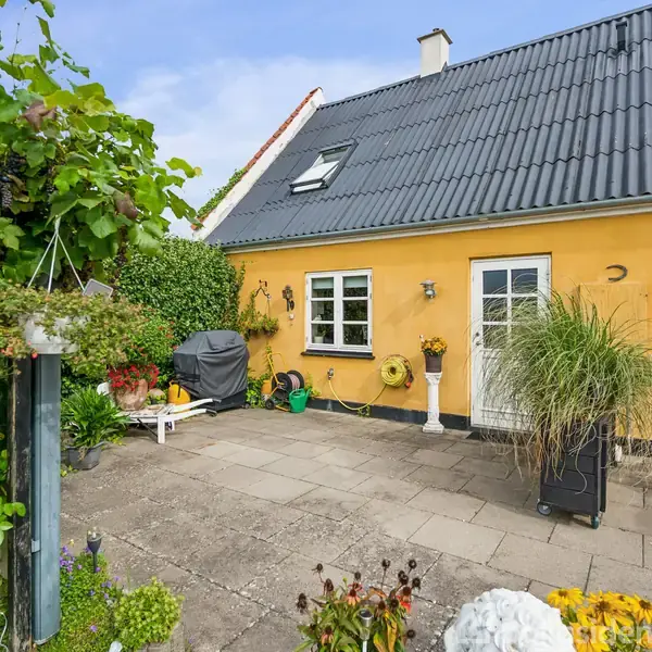 Gul husfacade med sorte tagsten, to døre og vindue, omgivet af grønne planter og blomsterkrukker på en flisebelagt terrasse. Solskinsdag med klar blå himmel i baggrunden.