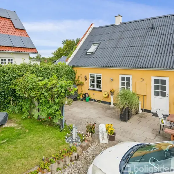 Gult hus med sorte tagsten har hvide døre og vinduer. Foran huset står en træbænk ved siden af blomster og en hvid bil i indkørslen.