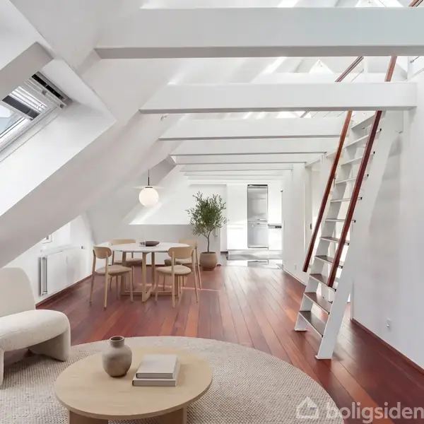 Et lyst, moderne loftsrum med skråvinduer. En stige leder til en hems. Rundt bord med stole står centralt, omgivet af minimalistisk indretning. Trægulve giver varme til rummet.
