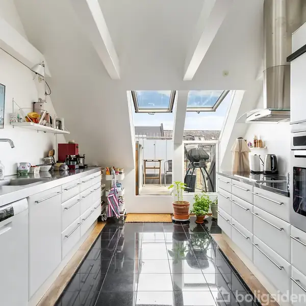Et moderne køkken med hvide skabe og sorte gulvfliser. Sollys strømmer ind gennem store ovenlysvinduer. Køkkenet er veludstyret med apparater og planter, og der er en dør til en terrasse.