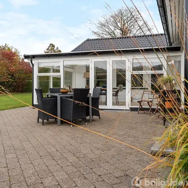 Terrassemøbler står på en flisebelagt patio foran et moderne anneks med vinduer. Omkring er en grøn have med buske og græsplæne, og himlen er let overskyet.