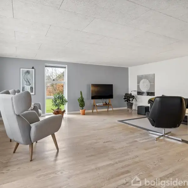 Stue med lyse trægulve indeholder grå lænestole, sort sofa, fladskærm på væg, planter, og stort vindue mod have. Maleri og lys i baggrunden.