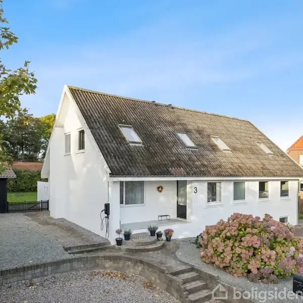 Hvidt hus med sadeltag og flere ovenlysvinduer, beliggende i en gruset have med buskads. Adressenummer 3 er synligt ved hoveddøren. En anden rød bygning ses i baggrunden.